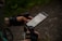 A person wearing fingerless gloves holds a smartphone displaying a map application. The person is also holding a walking pole, suggesting they might be hiking. The background is outdoors with a gravel path and greenery.
