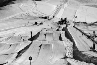 A snowy ski resort landscape with various slopes and paths. Skiers and snowboarders are visible on the runs, using the ski lift on the right. The scene captures the structured terrain with tracks and marks in the snow, and the recreational activity.