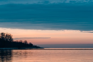 A serene landscape featuring a calm body of water reflecting the soft hues of a sunset. The sky transitions from a deep blue at the top to warm shades of orange and pink near the horizon. Silhouetted trees line the left side of the image, creating a peaceful and tranquil scene.