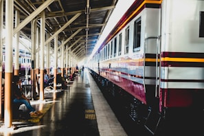 A long passenger train is parked at a railway station platform under a canopy. Several people are seated on benches along the platform, some with luggage. The station is brightly lit, with sunlight streaming through the gaps in the canopy.