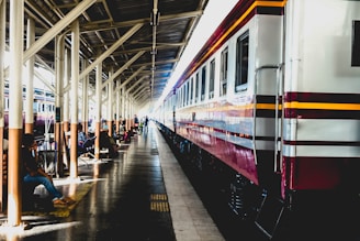 A long passenger train is parked at a railway station platform under a canopy. Several people are seated on benches along the platform, some with luggage. The station is brightly lit, with sunlight streaming through the gaps in the canopy.