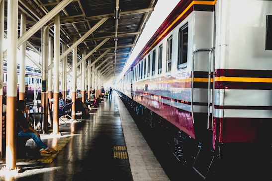 A long passenger train is parked at a railway station platform under a canopy. Several people are seated on benches along the platform, some with luggage. The station is brightly lit, with sunlight streaming through the gaps in the canopy.
