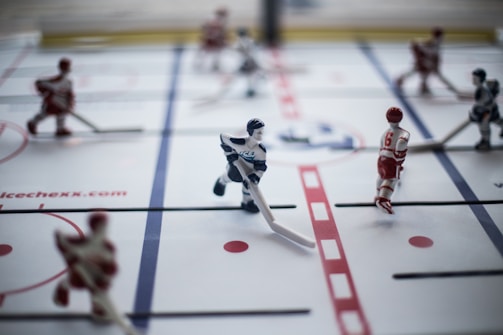 A close-up of a tabletop hockey game with plastic players mounted on rods. The players are positioned on a marked rink, with some wearing red uniforms and others in white. The surface includes lines mimicking a real hockey rink, with red and blue markings.