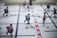 A close-up of a tabletop hockey game with plastic players mounted on rods. The players are positioned on a marked rink, with some wearing red uniforms and others in white. The surface includes lines mimicking a real hockey rink, with red and blue markings.