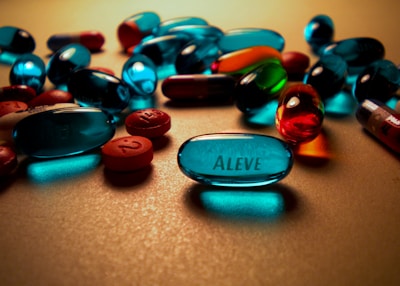 Various types of pills scattered across a surface, including several blue capsules with the label 'Aleve' visible. The lighting creates reflections and shadows that enhance the vibrant colors of the capsules.
