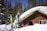 A wooden cabin is surrounded by a thick layer of snow, with a collection of skis and poles propped against the exterior. Tall pine trees are in the background, under a clear blue sky, creating a serene winter scene.