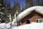 A wooden cabin is surrounded by a thick layer of snow, with a collection of skis and poles propped against the exterior. Tall pine trees are in the background, under a clear blue sky, creating a serene winter scene.
