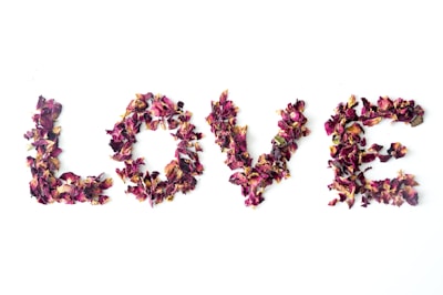 The word 'LOVE' is formed using dried rose petals arranged on a white background. The petals appear in shades of red, pink, and purple, creating a vibrant and textured appearance against the light backdrop.