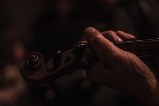 A hand gently holds the neck of a violin, with fingers poised delicately on the strings. The image is dimly lit, emphasizing the warmth and texture of the wood.