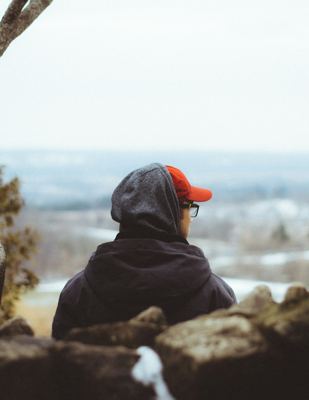 person in black hoodie and orange knit cap sitting on rock near body of water during