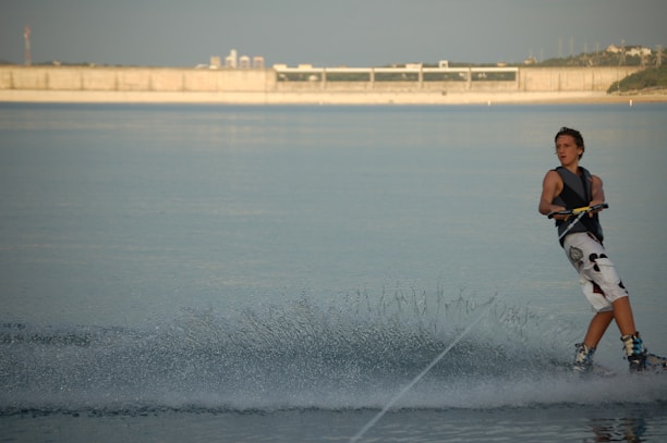 A person engaging in wakeboarding on a calm body of water with a concrete structure in the background. The rider is wearing a life jacket and maneuvering through the water, creating a trail of spray behind them.