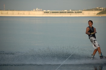 A person engaging in wakeboarding on a calm body of water with a concrete structure in the background. The rider is wearing a life jacket and maneuvering through the water, creating a trail of spray behind them.