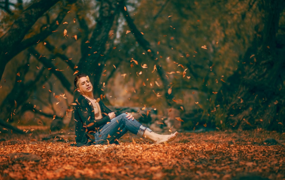 shallow focus photography of man sitting on brown dried leaves surrounded by trees during daytime