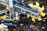 A detailed LEGO construction resembling a machine or vehicle with intricate mechanical parts is displayed. The build features a prominent circular component with yellow blades and tracks like those on construction equipment. Several LEGO figures are interacting with the structure, adding a sense of scale and activity. The setup is placed on a natural background with wood chips scattered around.