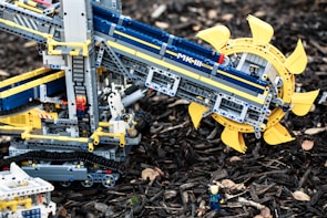 A detailed LEGO construction resembling a machine or vehicle with intricate mechanical parts is displayed. The build features a prominent circular component with yellow blades and tracks like those on construction equipment. Several LEGO figures are interacting with the structure, adding a sense of scale and activity. The setup is placed on a natural background with wood chips scattered around.