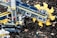 A detailed LEGO construction resembling a machine or vehicle with intricate mechanical parts is displayed. The build features a prominent circular component with yellow blades and tracks like those on construction equipment. Several LEGO figures are interacting with the structure, adding a sense of scale and activity. The setup is placed on a natural background with wood chips scattered around.
