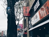 Buildings with signs in Japanese and English line a street, with a large tree in the foreground. A T-Mobile store is visible along with a Japanese restaurant named Kouraku. The scene is urban, with clear skies and some pedestrians.