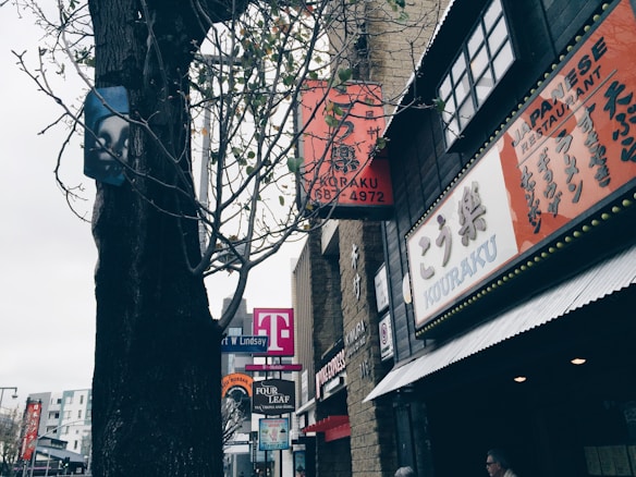 Buildings with signs in Japanese and English line a street, with a large tree in the foreground. A T-Mobile store is visible along with a Japanese restaurant named Kouraku. The scene is urban, with clear skies and some pedestrians.
