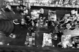A bustling bookshop with numerous people gathered around tables filled with books. The interior is spacious with a wooden floor and shelves packed with books are visible along the wall. The scene is viewed from above, capturing the activity of people browsing, reading, and interacting.