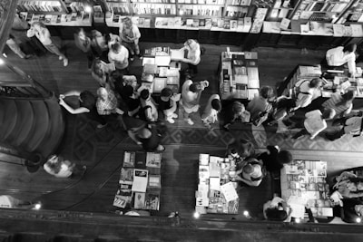 A bustling bookshop with numerous people gathered around tables filled with books. The interior is spacious with a wooden floor and shelves packed with books are visible along the wall. The scene is viewed from above, capturing the activity of people browsing, reading, and interacting.