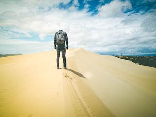 A person with a backpack is walking alone on smooth, golden sand dunes under a partly cloudy sky. Footprints trail behind, indicating a recent path across the gently curving landscape. Hills or mountains are faintly visible in the distance, providing a contrasting backdrop.