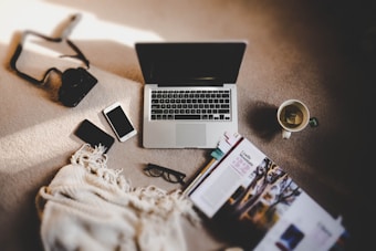A cozy flat lay featuring a laptop, two smartphones, a camera, a pair of glasses, a magazine, a hot beverage in a mug, and a fringed blanket spread out on a carpet. The items are arranged in a relaxed, casual manner, suggesting a comfortable work or leisure setting.