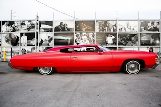 A sleek, low-rider red car is parked on a concrete surface, set against a backdrop of a wall featuring a collage of black and white historical and cultural photographs. The car has distinctive elongated bodywork and shiny chrome wheels, giving it a classic, vintage appeal.