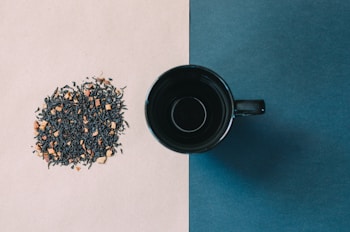 A black coffee cup sits on a background divided into two colors, beige on the left and dark blue on the right. On the beige side, there's a pile of loose leaf tea with visible dried fruits or flowers mixed in.