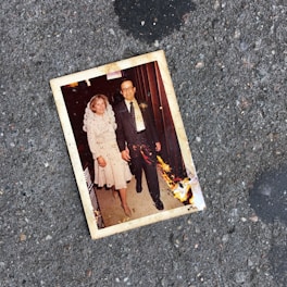 A photograph of an older couple holding hands is lying on a rough, gray pavement. The photograph has some burnt damage on its lower right corner. The couple appears well-dressed; the woman is wearing a light-colored dress with lace details, and the man is in a dark suit with a patterned tie.