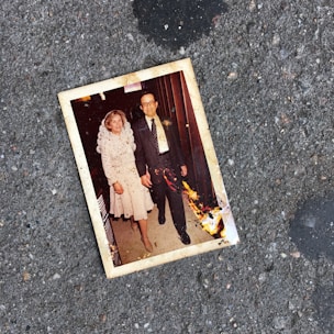 A photograph of an older couple holding hands is lying on a rough, gray pavement. The photograph has some burnt damage on its lower right corner. The couple appears well-dressed; the woman is wearing a light-colored dress with lace details, and the man is in a dark suit with a patterned tie.