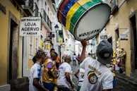 A vibrant street scene featuring a group of people, some wearing colorful shirts, participating in a lively parade or procession. One individual is prominently holding a large drum decorated with multicolored stripes. The narrow cobblestone street is flanked by buildings with rustic aesthetics, housing various shops indicated by signs in the Portuguese language.