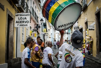 A vibrant street scene featuring a group of people, some wearing colorful shirts, participating in a lively parade or procession. One individual is prominently holding a large drum decorated with multicolored stripes. The narrow cobblestone street is flanked by buildings with rustic aesthetics, housing various shops indicated by signs in the Portuguese language.