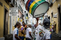 A vibrant street scene featuring a group of people, some wearing colorful shirts, participating in a lively parade or procession. One individual is prominently holding a large drum decorated with multicolored stripes. The narrow cobblestone street is flanked by buildings with rustic aesthetics, housing various shops indicated by signs in the Portuguese language.