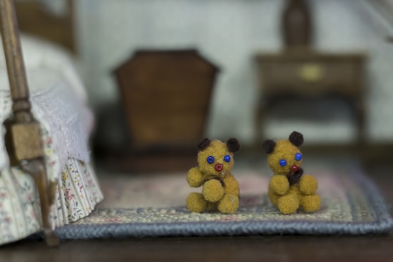 Two small, fuzzy teddy bears with blue eyes and red noses sit side by side on a patterned rug. The background shows parts of a cozy bedroom with a floral bedspread and a wooden dresser, slightly out of focus.