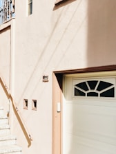 A portion of a building facade with a stucco texture, featuring a set of stairs leading up to a landing. The building is painted in a soft beige color. A garage door is present with a decorative window pattern at the top, accompanied by a small rectangular plaque above the door displaying the number 307 in dark lettering. There are three small square windows embedded in the wall next to the stairs.