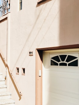 A portion of a building facade with a stucco texture, featuring a set of stairs leading up to a landing. The building is painted in a soft beige color. A garage door is present with a decorative window pattern at the top, accompanied by a small rectangular plaque above the door displaying the number 307 in dark lettering. There are three small square windows embedded in the wall next to the stairs.