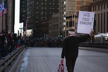 A man walks down an empty street between tall buildings, holding a sign that reads 'US AND EUROPE.' Flags and spectators line the side of the street. The setting suggests an urban environment, possibly during a demonstration or public event.