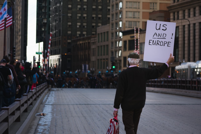 A man walks down an empty street between tall buildings, holding a sign that reads 'US AND EUROPE.' Flags and spectators line the side of the street. The setting suggests an urban environment, possibly during a demonstration or public event.