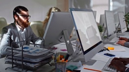 Several people are working at computers in an office environment. They are seated in a row, each focused on their own screen. The desk is equipped with office supplies, a document tray filled with papers, and various stationery. The lighting creates bright spots and soft shadows across the area.