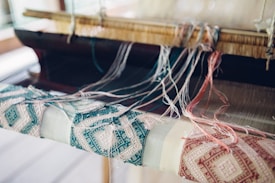 An intricate weaving setup with threads in various colors strewn across a loom, creating a detailed pattern on the fabric. The focus is on the woven textile, which features geometric designs in teal and beige tones, alongside loose threads and the wooden parts of the loom.