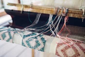 An intricate weaving setup with threads in various colors strewn across a loom, creating a detailed pattern on the fabric. The focus is on the woven textile, which features geometric designs in teal and beige tones, alongside loose threads and the wooden parts of the loom.