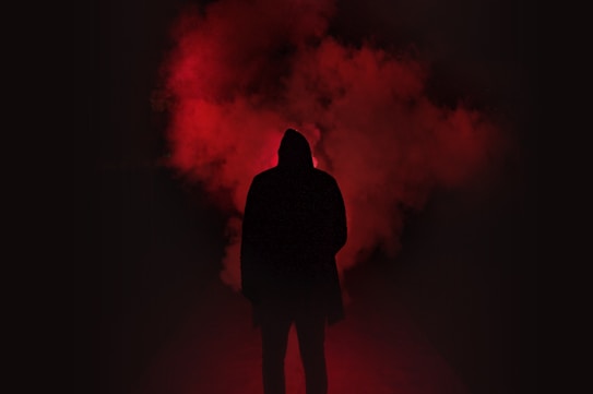 A silhouette of a person wearing a hooded jacket stands against a backdrop of dense red smoke, creating a mysterious and intense atmosphere. The dark surroundings contrast sharply with the vivid red, enhancing the dramatic effect.