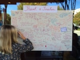 A large poster filled with handwritten thank-you notes dedicated to teachers, written in various colors such as red, blue, and purple. A child with blonde hair is writing a message on the poster, focusing intently. The poster is displayed on a window, and a reflection shows trees and a building outside.