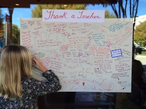 A large poster filled with handwritten thank-you notes dedicated to teachers, written in various colors such as red, blue, and purple. A child with blonde hair is writing a message on the poster, focusing intently. The poster is displayed on a window, and a reflection shows trees and a building outside.