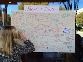 A large poster filled with handwritten thank-you notes dedicated to teachers, written in various colors such as red, blue, and purple. A child with blonde hair is writing a message on the poster, focusing intently. The poster is displayed on a window, and a reflection shows trees and a building outside.