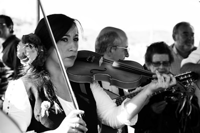 A group of musicians is gathered, with a woman in traditional clothing playing a violin. Her hair is adorned with a flower, and she exhibits a focused expression. Other musicians are in the background, playing string instruments.