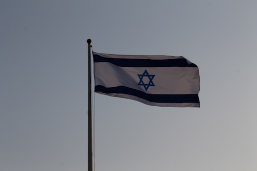 A flag featuring a blue Star of David centered between two horizontal blue stripes on a white background, fluttering against a clear sky.