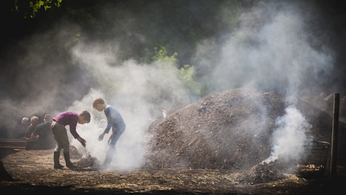 Two children are crouching near a large smoldering pile of earth or charcoal, surrounded by a cloud of smoke. They appear to be engaged in a task, possibly related to making charcoal. In the background, other people are watching or resting near a wooden bench. Sunlight filters through the trees, casting a soft light over the scene.