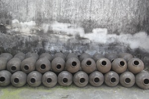A line of spherical earthenware pots is arranged on the ground in front of a grey, worn concrete wall. There are two rows of these pots, each displaying a textured surface, and they appear to be empty, with their openings facing outward.
