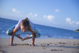 A person performing a yoga pose on a sandy beach with the ocean and a clear blue sky in the background. The person is balancing on their hands with their legs folded underneath them, demonstrating strength and balance.